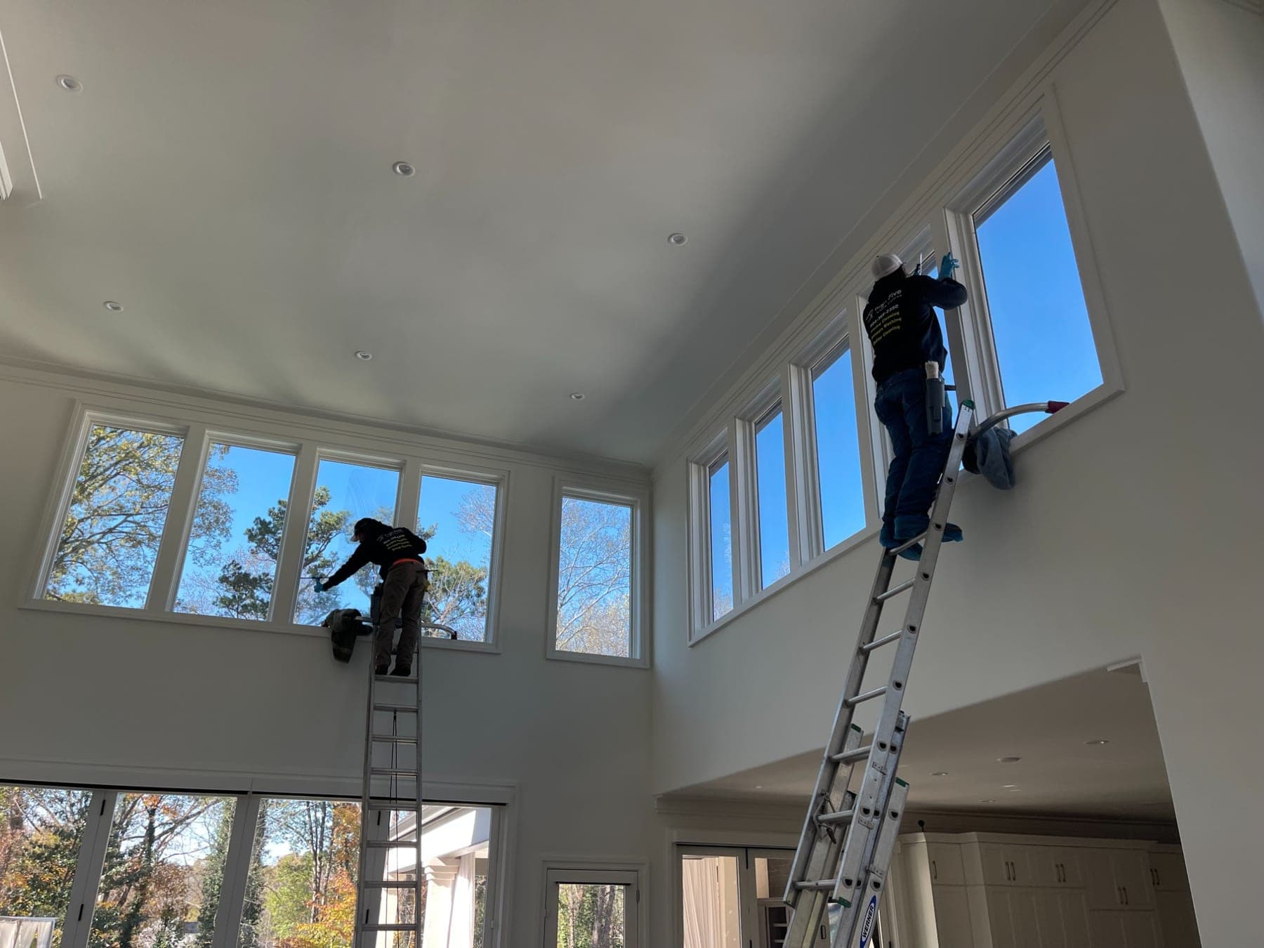 Two High Five cleaners on ladders, washing tall interior windows in a two-story room