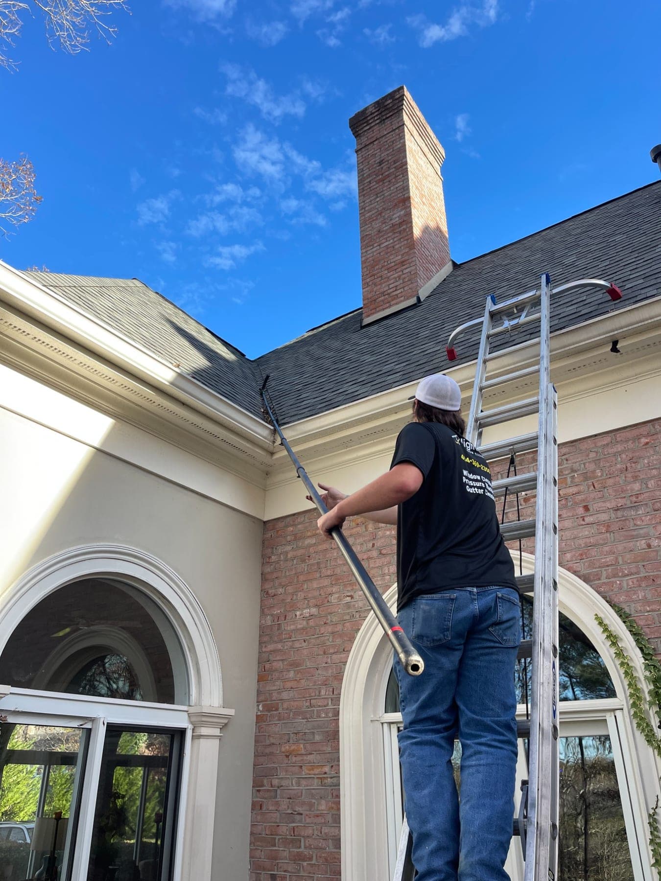 A High Five cleaner on a ladder clearing a roof valley above the gutter beside a brick chimney