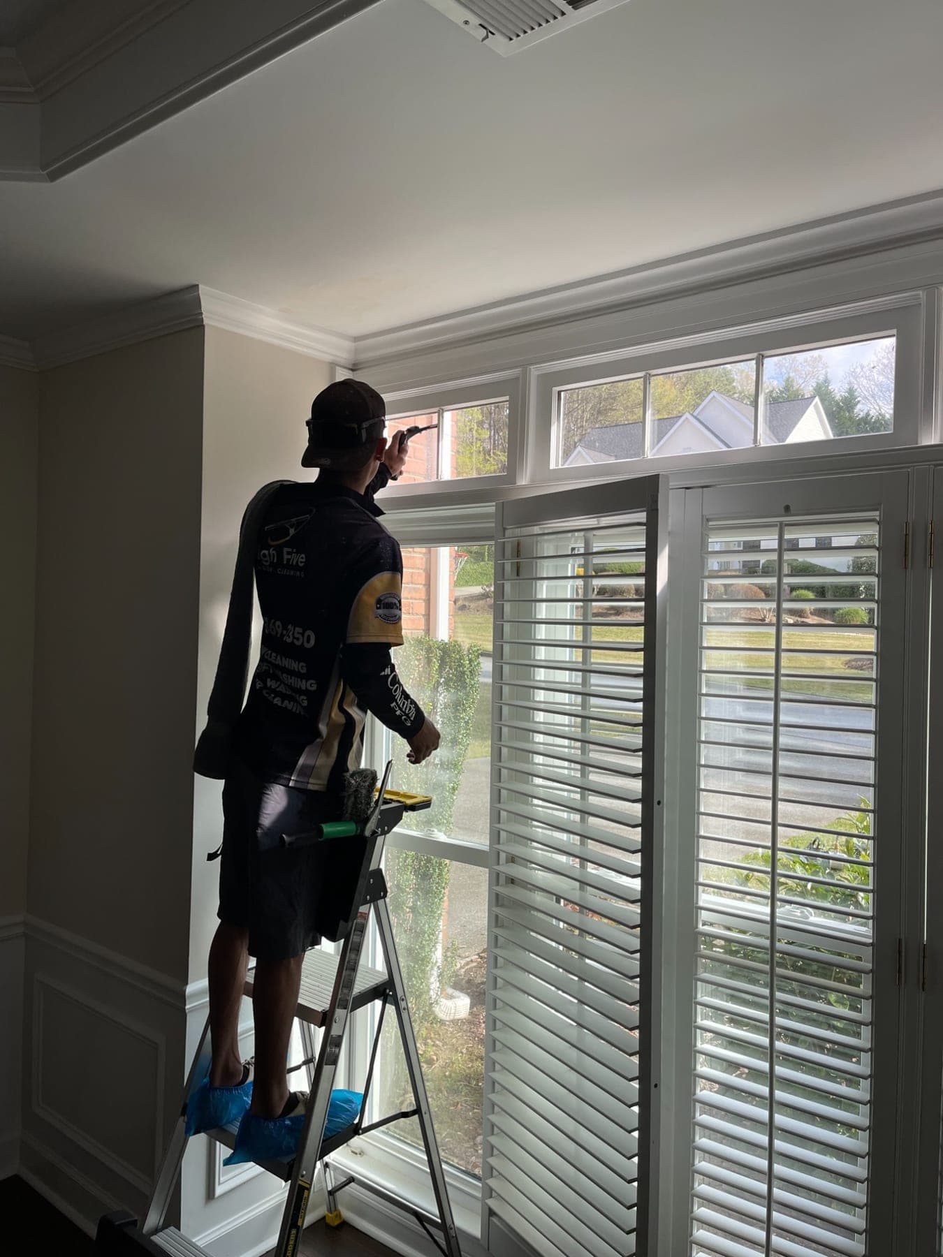 A High Five cleaner on a ladder inside a home, wearing blue shoe covers while cleaning transom windows